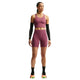 En person står med ansigtet vendt fremad foran en hvid baggrund og har Nikes Dame High-Waisted 7″ løbeshorts med lommer i farven Pink Smoke/Reflective Silver på, en matchende lilla sports-BH, sorte armvarmere, sorte sokker og flerfarvede sportssko.