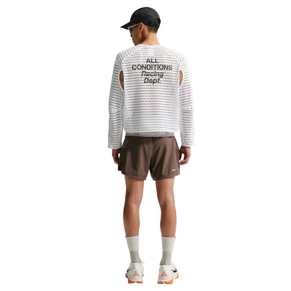 En person står med ryggen til og har en Nike ACG Radical AirFlow NXT langærmet løbetrøje til mænd i farvekombinationen hvid/antracit på, kombineret med brune shorts, hvide sokker, sneakers og en sort kasket. Den enkle baggrund antyder inspiration fra trail-løb.