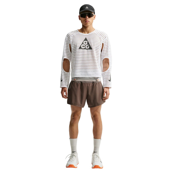 En person står foran kameraet iført solbriller, en sort kasket, brune shorts, lange beige sokker samt en Nike ACG Radical AirFlow NXT langærmet løbetrøje til mænd i farvekombinationen hvid/antracit med udskæringer ved albuerne og Nike trail-løbesko med orange såler.