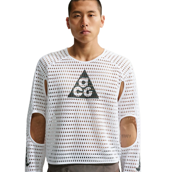 En person har Nike ACG Radical AirFlow NXT langærmet løbetrøje til mænd i farvekombinationen hvid/antracit på. Trøjen er fremstillet af hvidt mesh-stof med runde huller og udskæringer ved ærmerne samt et sort trekantet logo på brystet på en ensfarvet baggrund.