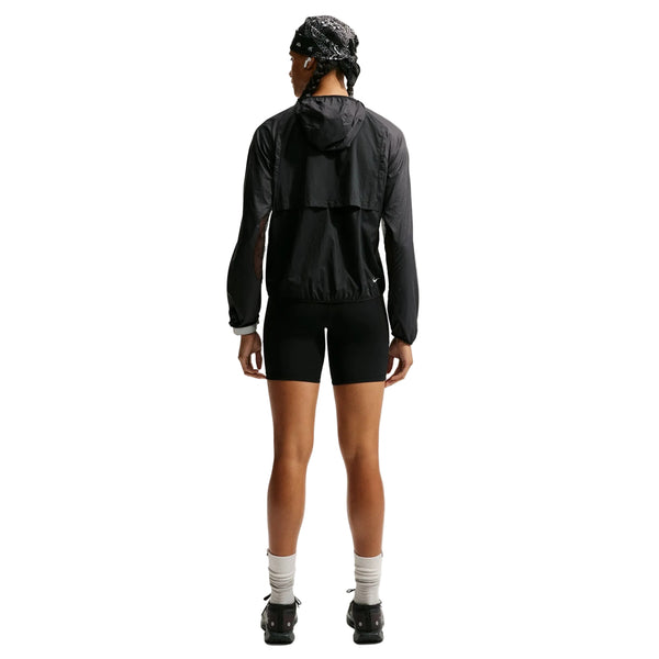 En person står med ryggen til og har en Nike ACG »Aireez« UV-beskyttelsesjakke til kvinder i farvekombinationen sort/sort/Summit White på, kombineret med sorte shorts, hvide sokker, sorte sko og et mønstret bandana. Baggrunden er hvid.