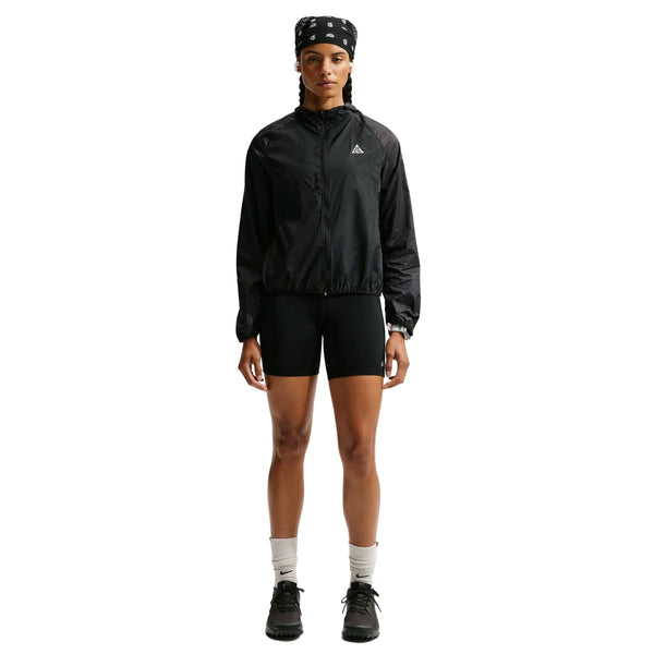 En person står med ansigtet vendt fremad iført en Nike ACG »Aireez« UV-beskyttelsesjakke til kvinder i farvekombinationen sort/sort/Summit White, samt sorte cykelshorts, sorte sko, hvide sokker og et sort bandana. Baggrunden er hvid og ryddelig.