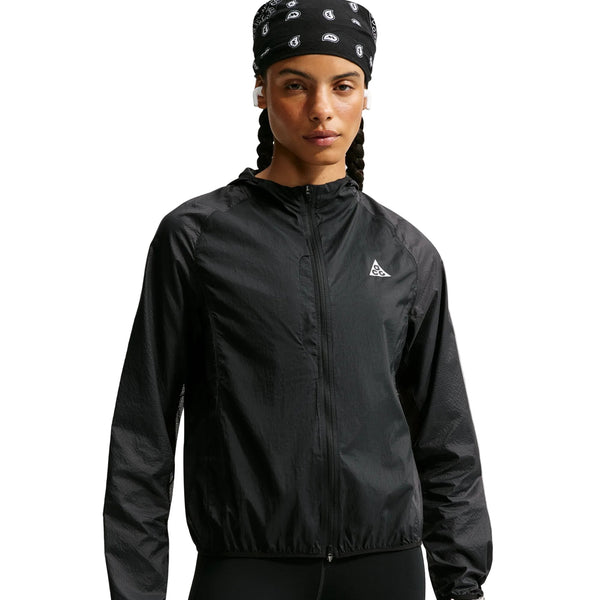 En kvinde står med ansigtet vendt mod kameraet foran en hvid baggrund iført Nike ACG »Aireez« UV Protection Jacket i farvekombinationen sort/sort/summit white – en elegant vindjakke med lynlås, der har et lille hvidt logo på brystet. Kombineret med en sort bandana med hvide mønstre og trådløse øretelefoner forener denne vandafvisende løbejakke stil og funktionalitet, hvilket gør den til et fremragende valg for kvinder, der søger pålidelig vejrbeskyttelse under udendørsaktiviteter.