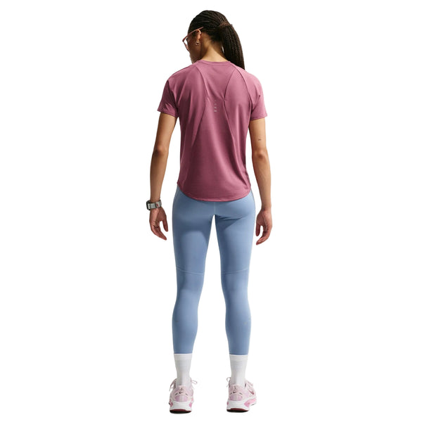 En person står med ryggen til kameraet og har en Nike Dri-FIT-løbetrøje til kvinder i farven Pink Smoke/Reflective Silver på, som er svedtransporterende, samt lyseblå leggings, hvide sokker, lyserøde sneakers, et ur og briller. Håret er sat op i fletninger. Baggrunden er ensfarvet hvid.