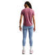 En person står med ryggen til kameraet og har en Nike Dri-FIT-løbetrøje til kvinder i farven Pink Smoke/Reflective Silver på, som er svedtransporterende, samt lyseblå leggings, hvide sokker, lyserøde sneakers, et ur og briller. Håret er sat op i fletninger. Baggrunden er ensfarvet hvid.