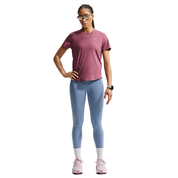 En kvinde står med ansigtet vendt fremad og den ene hånd på hoften foran en hvid baggrund. Hun har en Nike Dri-FIT løbetrøje til kvinder i farven Pink Smoke/Reflective Silver på, kombineret med lyseblå leggings, hvide sokker, lyserøde sneakers, store solbriller og et smartwatch. Den svedtransporterende Dri-FIT Running Top fra Nike fremhæver hendes outfit med både stil og komfort.