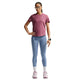 En kvinde står med ansigtet vendt fremad og den ene hånd på hoften foran en hvid baggrund. Hun har en Nike Dri-FIT løbetrøje til kvinder i farven Pink Smoke/Reflective Silver på, kombineret med lyseblå leggings, hvide sokker, lyserøde sneakers, store solbriller og et smartwatch. Den svedtransporterende Dri-FIT Running Top fra Nike fremhæver hendes outfit med både stil og komfort.