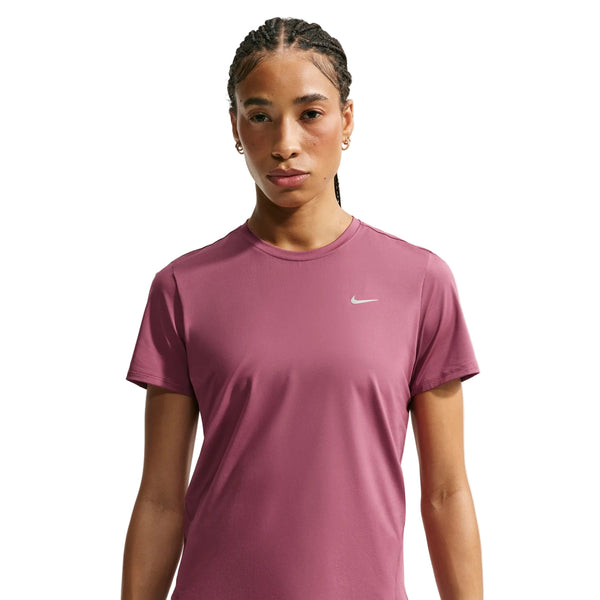 En kvinde med flettet hår har en lyserød Nike Dri-FIT-løbetrøje til kvinder på, der har reflekterende sølvdetaljer og svedtransporterende egenskaber, samt små ringøreringe. Hun står foran en ensfarvet hvid baggrund og ser direkte ind i kameraet med et neutralt udtryk.