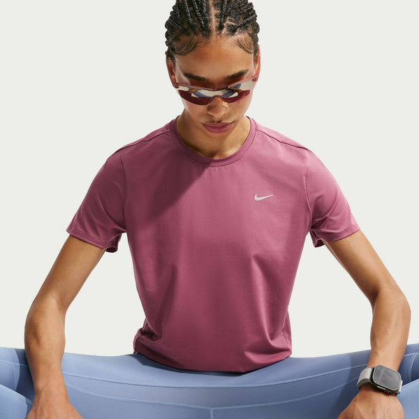 En kvinde iført Nike Dri-FIT løbetrøje, Dame, Pink Smoke/Reflective Silver, solbriller og et Apple Watch kigger ned, mens hun sidder foran en lys baggrund. Hendes hår er flettet, og hendes arme hviler på et blåt stykke stof. Den svedtransporterende Nike-løbetrøje sikrer komfort – også i afslappede øjeblikke.