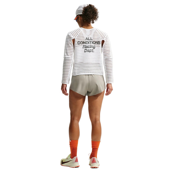 En person står med ryggen til og er iført en hvid langærmet netbluse med teksten »ALL CONDITIONS Racing Dept« på ryggen, Nike ACG »Trailwind«-shorts til kvinder i farven Light Army/White, orange sokker, hvide og røde sko samt en hvid kasket med skygge. Baggrunden er ensfarvet hvid.