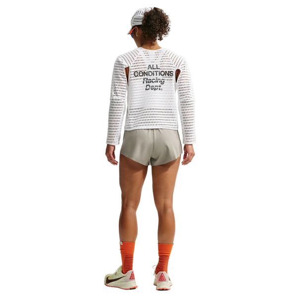 En person står med ryggen til og er iført en hvid langærmet netbluse med teksten »ALL CONDITIONS Racing Dept« på ryggen, Nike ACG »Trailwind«-shorts til kvinder i farven Light Army/White, orange sokker, hvide og røde sko samt en hvid kasket med skygge. Baggrunden er ensfarvet hvid.