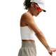 En kvinde står foran en hvid baggrund iført en hvid crop top, Nike ACG »Trailwind«-shorts til kvinder i farven Light Army/White, en hvid kasket med huller og orange solbriller med reflekser. Hun har en tatovering på venstre underarm og kigger nedad.