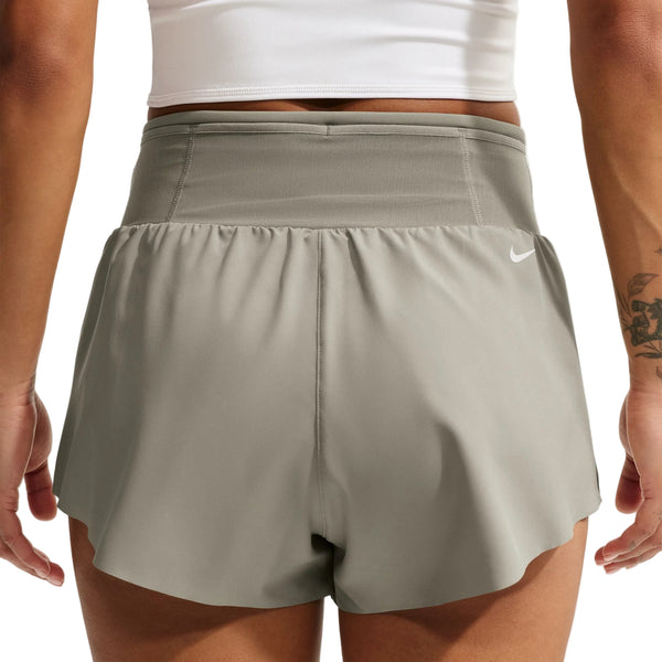 Der ses en person set bagfra, iført en lys militæruniform og hvide Nike ACG »Trailwind«-shorts til kvinder samt en hvid crop-top. En del af personens armtatovering er synlig, og hænderne hviler afslappet langs siden.