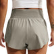 Der ses en person set bagfra, iført en lys militæruniform og hvide Nike ACG »Trailwind«-shorts til kvinder samt en hvid crop-top. En del af personens armtatovering er synlig, og hænderne hviler afslappet langs siden.