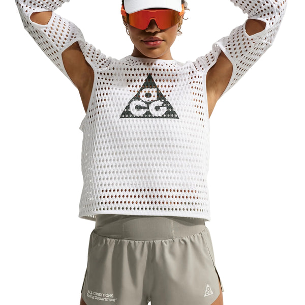 En person har en hvid langærmet trøje i mesh med et trekantet logo på, kombineret med Nike ACG »Trailwind«-shorts til kvinder i farven Light Army/White, hvor der er en lille tekst nær nederkanten. Personen har desuden en hvid kasket og røde solbriller med skygge på, mens vedkommende poserer med armene løftet over hovedet.