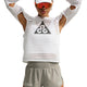 En person har en hvid langærmet trøje i mesh med et trekantet logo på, kombineret med Nike ACG »Trailwind«-shorts til kvinder i farven Light Army/White, hvor der er en lille tekst nær nederkanten. Personen har desuden en hvid kasket og røde solbriller med skygge på, mens vedkommende poserer med armene løftet over hovedet.