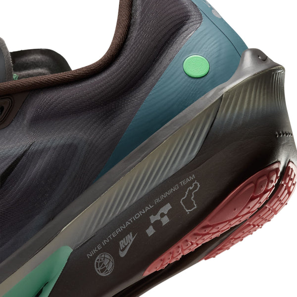 Nærbillede af hælen på Nike Zoom Fly 6 Herre i farvekombinationen Baroque Brown/Black-Smoke Grey-Ironstone, der fremhæver den strukturerede ydersål, teksten »Nike International Running Team« og forskellige logoer. Skoen har et elegant design i kulfiber med mørke og blågrønne nuancer, en grøn prikdetalje og synlige brune snørebånd.