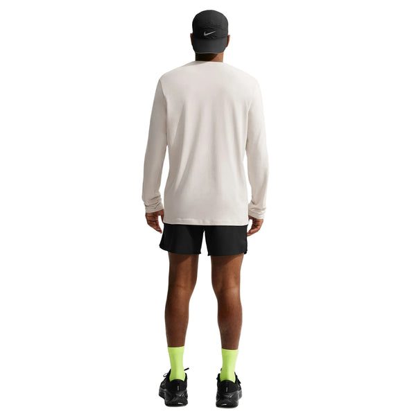 En mand står med ryggen til kameraet. Han har en sort kasket på, en Nike Dri-FIT langærmet løbetrøje til mænd i farven »Light Bone«, sorte shorts, neongrønne sokker og sorte sko. Baggrunden er hvid og ryddelig.