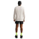 En mand står med ryggen til kameraet. Han har en sort kasket på, en Nike Dri-FIT langærmet løbetrøje til mænd i farven »Light Bone«, sorte shorts, neongrønne sokker og sorte sko. Baggrunden er hvid og ryddelig.
