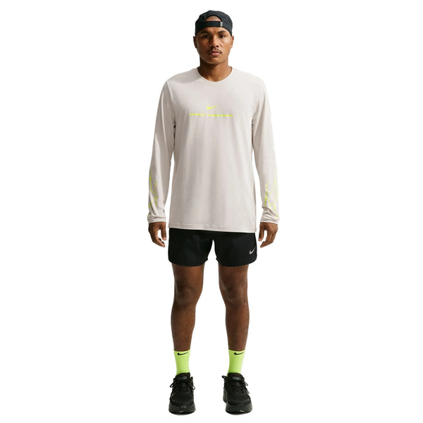 En mand står med ansigtet vendt fremad iført en Nike Dri-FIT langærmet løbetrøje, Herre, i farven Light Bone, samt sorte shorts, neongule sokker, sorte sneakers og en sort kasket, der er vendt bagud. Baggrunden er ensfarvet hvid.