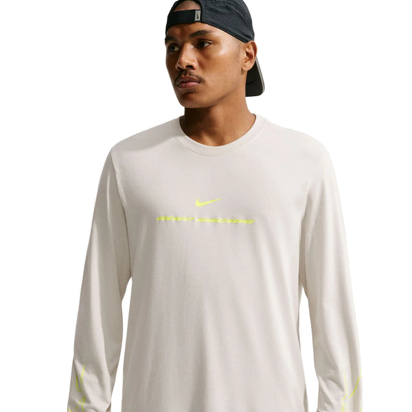 En person iført en Nike Dri-FIT langærmet løbetrøje i farven Light Bone og en sort kasket, der sidder baglæns, stående foran en ensfarvet hvid baggrund.