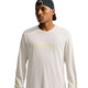 En person iført en Nike Dri-FIT langærmet løbetrøje i farven Light Bone og en sort kasket, der sidder baglæns, stående foran en ensfarvet hvid baggrund.