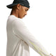 En person iført en sort kasket, der sidder baglæns, og en lys Nike Dri-FIT langærmet løbetrøje, Herre, Light Bone, står i profil foran en ensfarvet baggrund og ser ned med armene strakt fremad. Der ses neongule detaljer på det ene ærme af Nike-løbetrøjen.