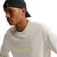 En person har en Nike Dri-FIT langærmet løbetrøje til kvinder i farven Light Bone på, med et neongult Nike-logo og tekst. Personen har desuden en sort kasket på bagvendt og står vendt mod venstre foran en ensfarvet hvid baggrund.