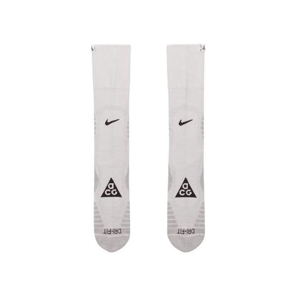 Nike ACG Crew Socks, Unisex i Summit White/Lt Smoke Grey, har sorte Swoosh-logoer i toppen og ACG-trekanter med DRI-FIT-tekst på foden, vist på en almindelig hvid baggrund. Fås i størrelserne S, M og L.