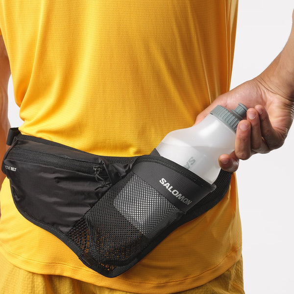 En person i en gul trøje lægger en Salomon Active Belt 3D-flaske, unisex, sort, ned i netlommen på sit sorte drikkebælte.