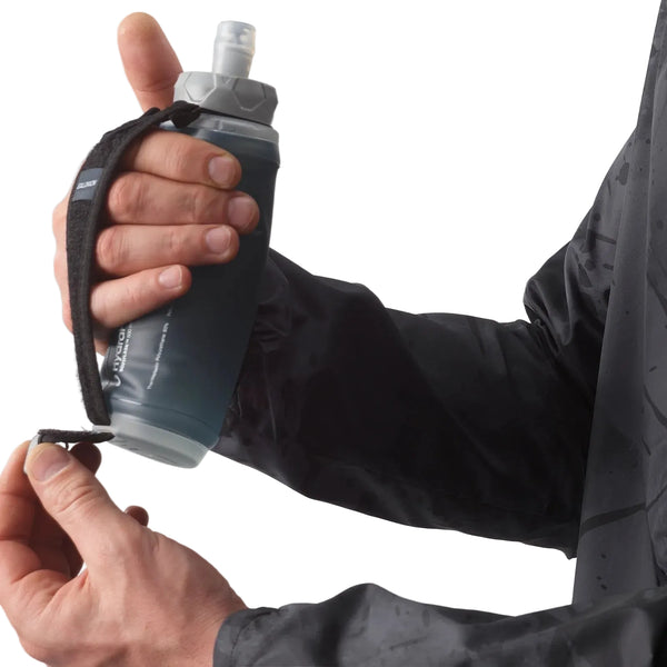 En person i en mørk jakke holder en Salomon Active Handheld-flaske (unisex, sort/skifergrå) med en justerbar rem – den ene hånd justerer remmen, mens den anden holder fast i flasken, klar til en løbetur.