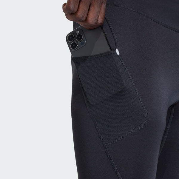 En person lægger en smartphone i netlommen på siden af Adidas Adi365 Adaptive Running Half Tights, Herre, Sort foran en ensfarvet, lys baggrund.