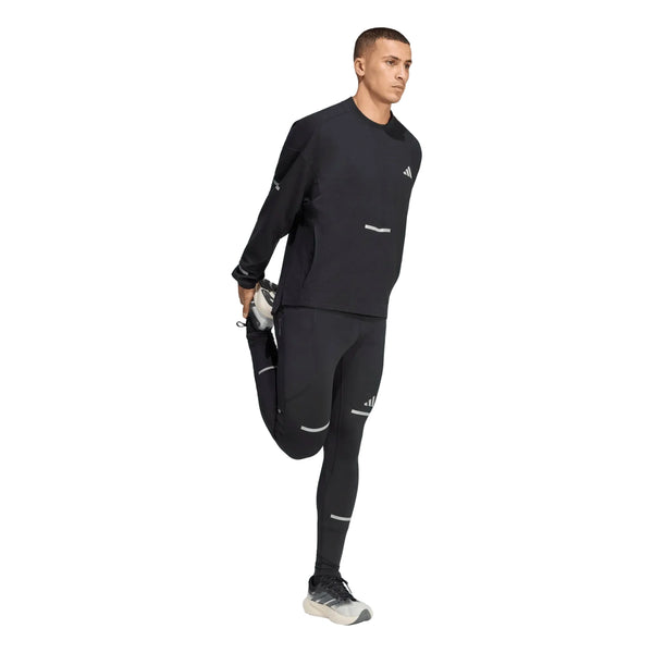 En person, der har en sort Adi365 Climawarm+ løbetrøje med rund hals og leggings på, udfører en stående strækøvelse for lårmusklerne.