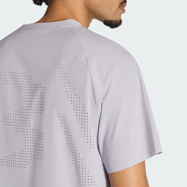 Der ses en person set bagfra og fra siden iført en Adidas Adi365 Running Aspyre T-shirt, Herre, Glogry, med små perforerede huller i et mønster på den øverste del af ryggen. Baggrunden er ensfarvet og lys.