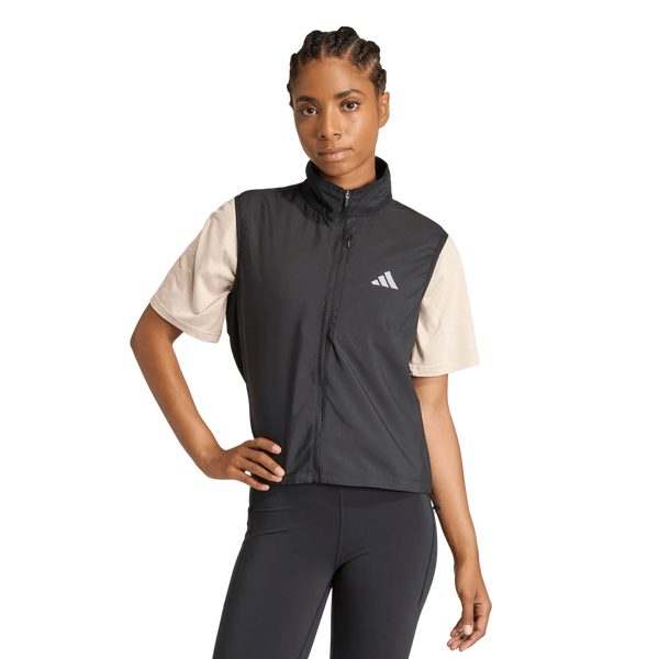 En person står med den ene hånd på hoften og er iført en lys beige T-shirt, en sort Adidas Adi365 Running Essentials-vest til kvinder med et hvidt logo på brystet samt sorte leggings på en ensfarvet hvid baggrund.