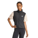 En person står med den ene hånd på hoften og er iført en lys beige T-shirt, en sort Adidas Adi365 Running Essentials-vest til kvinder med et hvidt logo på brystet samt sorte leggings på en ensfarvet hvid baggrund.