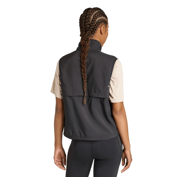 På billedet ses en person set bagfra med langt, flettet hår, iført en beige kortærmet bluse, sorte leggings og Adidas Adi365 Running Essentials-vest til kvinder i sort på en ensfarvet hvid baggrund.