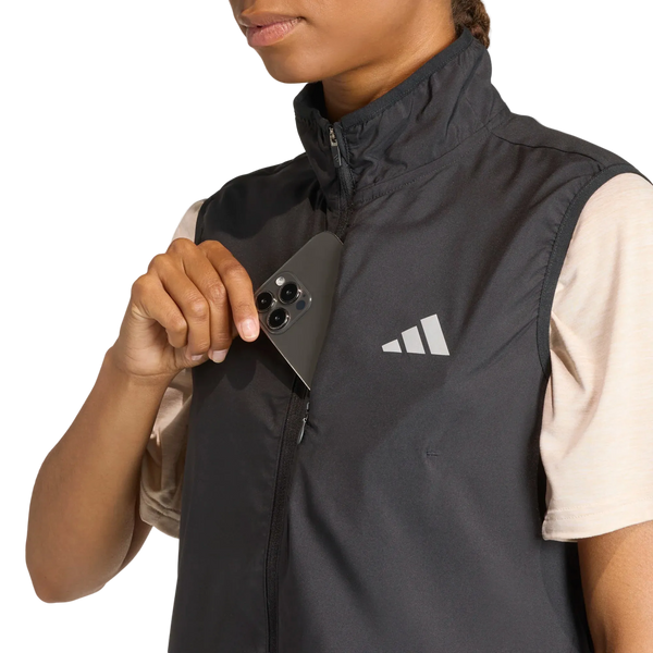 En kvinde iført en sort Adidas Adi365 Running Essentials-vest med et hvidt logo lægger en sort smartphone i brystlommen; hun har en beige kortærmet bluse på under vesten.