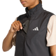 En kvinde iført en sort Adidas Adi365 Running Essentials-vest med et hvidt logo lægger en sort smartphone i brystlommen; hun har en beige kortærmet bluse på under vesten.