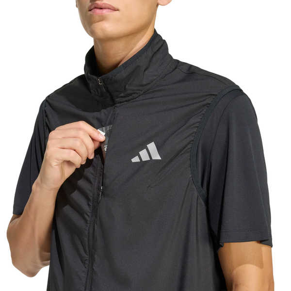 Herre har en sort Adi365 Running Essentials-vest fra Adidas på – en kortærmet sportsvest, der er delvist åbnet, så det karakteristiske hvide adidas-logo på brystet fremstår tydeligt mod en ensfarvet lys baggrund.
