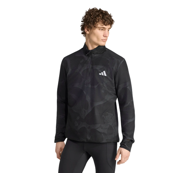 En person med krøllet hår står foran en ensfarvet hvid baggrund iført en Adidas Adi365 Running Half Zip langærmet trøje til mænd i farven Black/Grefiv, der har et hvidt adidas-logo på brystet, samt matchende sorte leggings.