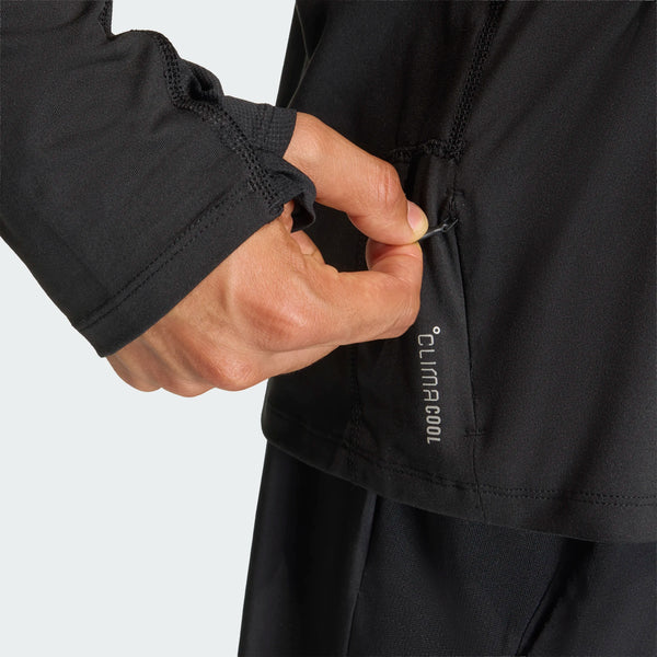 En person, der bærer en sort Adidas Adi365 Running Iconic Half Zip-jakke til mænd, bruger sin højre hånd til at lukke lynlåsen på en lille lomme på jakken, hvor der står »CLIMACOOL« tæt ved lynlåsen.