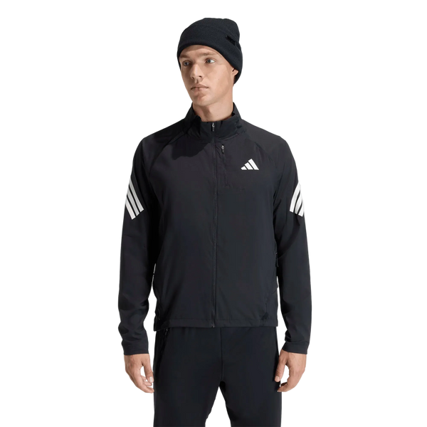 En mand står foran en ensfarvet hvid baggrund iført Adidas Adi365 Running Iconic-jakken, herre, sort, som er prydet med karakteristiske hvide Adidas-logoer og striber på ærmerne. Han kombinerer denne løbejakke med sorte bukser og en sort strikket hue, hvilket skaber et sporty og samtidig klassisk look, der forener både stil og komfort.