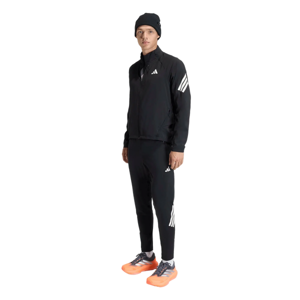 En person står med ansigtet vendt fremad foran en ensfarvet hvid baggrund og er iført en sort Adidas Adi365 Running Iconic-jakke (herre) med hvide Adidas-logoer og -striber, en sort hue samt lysorange og grå løbesko.