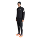 En person står med ansigtet vendt fremad foran en ensfarvet hvid baggrund og er iført en sort Adidas Adi365 Running Iconic-jakke (herre) med hvide Adidas-logoer og -striber, en sort hue samt lysorange og grå løbesko.