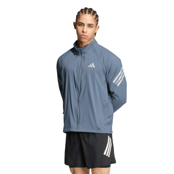 En person med flettet hår har en blå Adidas Adi365 Running Iconic-jakke, Herre, Onix/Refsil, med hvide striber på ærmerne og sorte shorts med hvide striber på, og står foran en ensfarvet hvid baggrund.