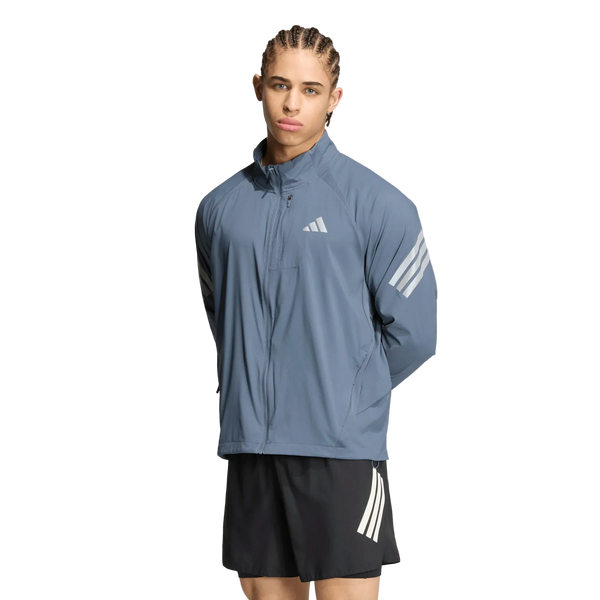 En person med flettet hår har en blå Adidas Adi365 Running Iconic-jakke, Herre, Onix/Refsil, med hvide striber på ærmerne og sorte shorts med hvide striber på, og står foran en ensfarvet hvid baggrund.