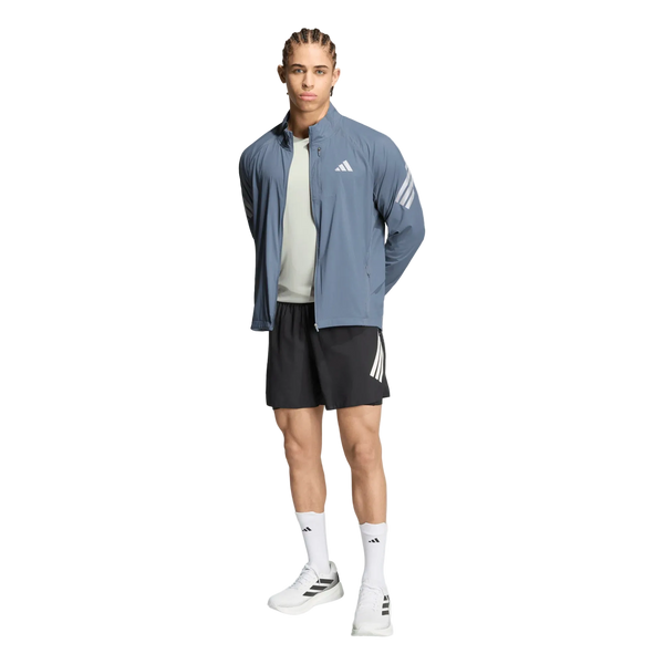 En person står med ansigtet vendt fremad og har en Adidas Adi365 Running Iconic-jakke til mænd i farven Onix/Refsil på – en blå løbejakke til mænd – kombineret med en lysegrå skjorte, sorte shorts, hvide strømper og hvide sportssko med sorte detaljer. Baggrunden er helt hvid.