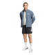 En person står med ansigtet vendt fremad og har en Adidas Adi365 Running Iconic-jakke til mænd i farven Onix/Refsil på – en blå løbejakke til mænd – kombineret med en lysegrå skjorte, sorte shorts, hvide strømper og hvide sportssko med sorte detaljer. Baggrunden er helt hvid.