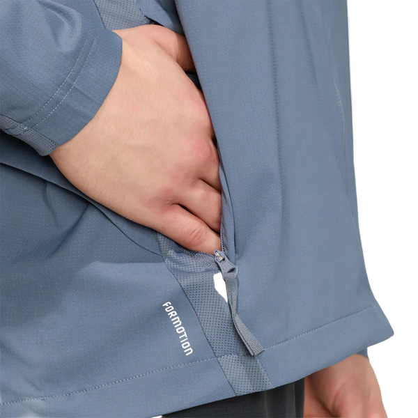 En persons venstre hånd stikker delvist ind i lommen med lynlås på en lyseblå Adidas Adi365 Running Iconic-jakke, Herre, Onix/Refsil. Ordet FORMATION er trykt tæt på jakkens nederste kant, og jakken har det slanke design, der er karakteristisk for Adidas.